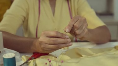 Asyalı kadın iğneyi ipliğe geçiriyordu. Stüdyosunda çalışan terzi, elinde odaklanmış dikiş iğnesine iplik koyuyor..
