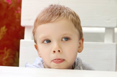 Restoran terasında oturan ve dilini gösteren komik çocuk. Arka planda çiçekler. Komik ekşi, beyaz saçlı ve mavi gözlü bir çocuk..