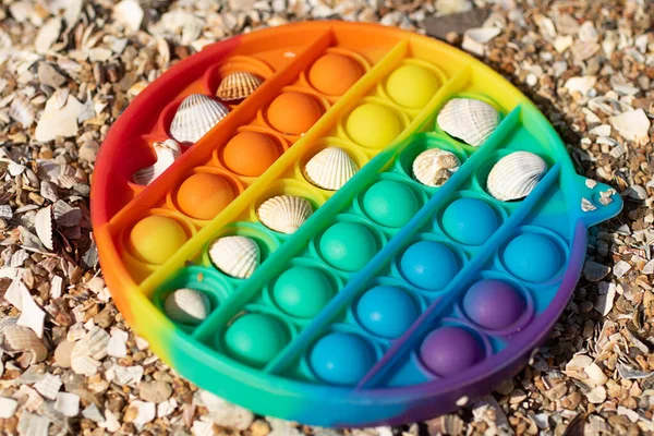 Nuevo juguete de silicona pop it en la playa con conchas en él, concepto creativo. Juguete antiestrés para niños y adultos. Colorear arco iris. — Foto de Stock