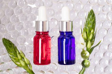 Kırmızı ve mavi camlardan yapılmış kozmetik şişeleri. Etraflarında şeffaf hidrojel topları. Çerçeve gibi beyaz çiçekli yeşil dallar..