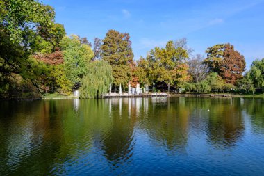 Romanya 'nın başkenti Bükreş' teki Carol Park 'ta, güneşli bir sonbahar gününde, göl kenarındaki büyük yeşil ve sarı ağaçların bulunduğu manzara