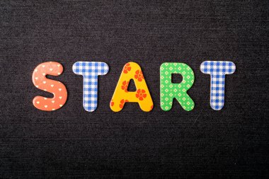 Başlangıç sözcüğünün bulunduğu kart, mesaj olarak kullanılabilecek koyu renkli siyah tekstil materyali üzerinde karışık renkli ahşap harflerden yapılmış.