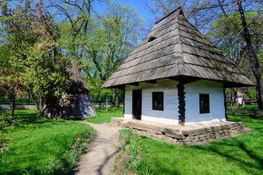 Bükreş, Romanya - 25 Nisan 2021: Eski geleneksel Romen evi güneşli bir bahar gününde Herastrau Park 'taki Dimitrie Gusti Ulusal Köy Müzesi' nde çok sayıda eski ağaç ve yeşil çimlerle çevrili