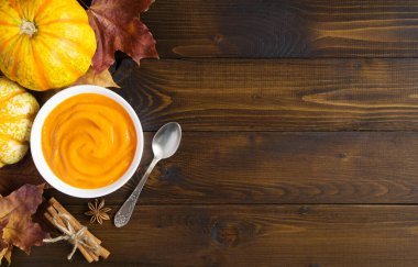 Beyaz kasede balkabağı kremalı çorba, sonbahar yaprakları, tarçın çubukları, kahverengi ahşap arka planda dekoratif balkabakları. Şükran Günü konsepti. Boşluğu kopyala. Üst görünüm.