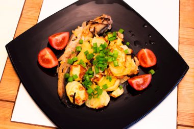 Patates domatesiyle süslenmiş pollock balığı. Siyah kare tabakta yeşillikler.