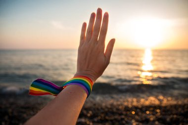 Bir gökkuşağı kurdelesiyle el, arka plandaki LGBT sembolü güneş ışınlarından geçen gün batımı.