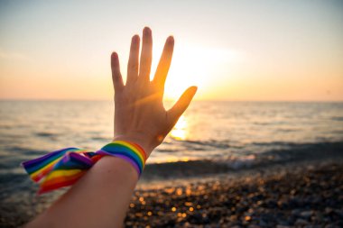 Bir gökkuşağı kurdelesiyle el, arka plandaki LGBT sembolü güneş ışınlarından geçen gün batımı.