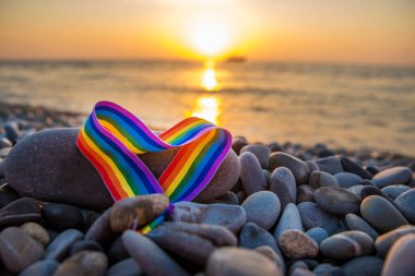 Kalp şeklinde gökkuşağı kurdelesi. Gün batımında deniz kenarındaki taşların üzerindeki LGBT sembolü.