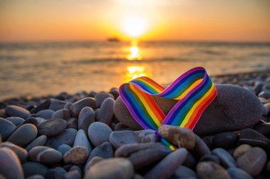 Kalp şeklinde gökkuşağı kurdelesi. Gün batımında deniz kenarındaki taşların üzerindeki LGBT sembolü.