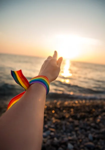 Hand with a rainbow ribbon - a symbol of LGBT on the background of a sea sunset through the sun\'s rays