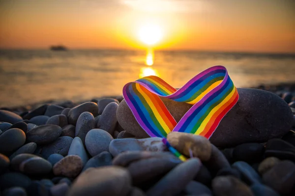 Kalp şeklinde gökkuşağı kurdelesi. Gün batımında deniz kenarındaki taşların üzerindeki LGBT sembolü.