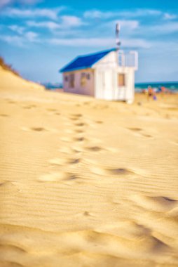 The road along the sandy beach to the blurry out of focus white summer beach house with a blue roof on the beach by the sea. Summer landscape