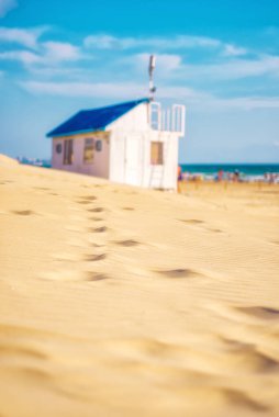 The road along the sandy beach to the blurry out of focus white summer beach house with a blue roof on the beach by the sea. Summer landscape