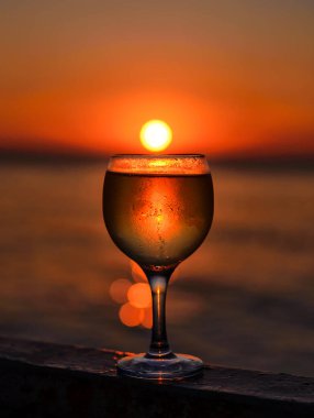 Güneşin renkli bir günbatımında kumsalda battığı bir bardak beyaz şarap. Bardakta güneş