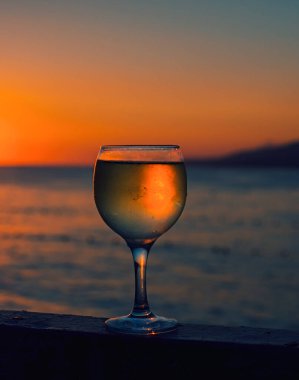 Güneşin renkli bir günbatımında kumsalda battığı bir bardak beyaz şarap. Bardakta güneş