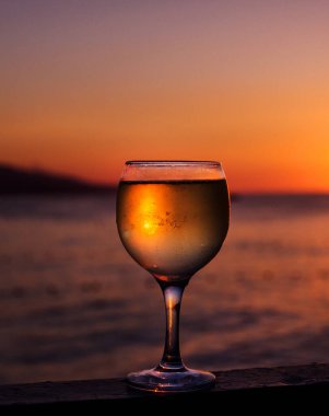 Güneşin renkli bir günbatımında kumsalda battığı bir bardak beyaz şarap. Bardakta güneş