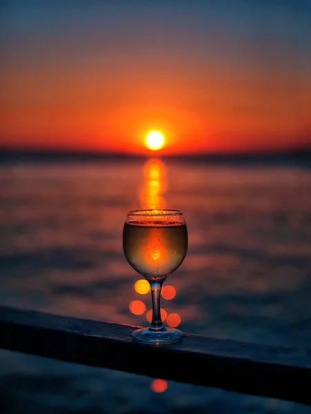 Güneşin renkli bir günbatımında kumsalda battığı bir bardak beyaz şarap. Bardakta güneş