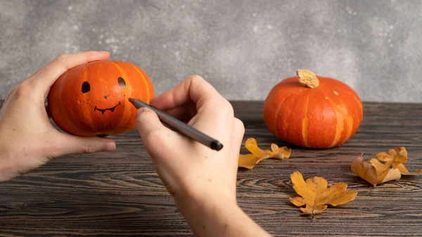 Womans hands drawing cute and funny Jack face on little pumpkin with black marker pen for Halloween party.
