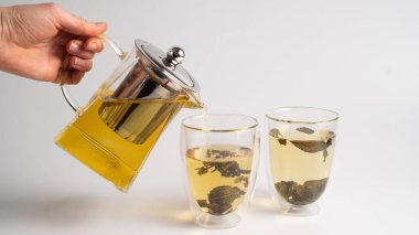 El, şeffaf bir çaydanlıktan cam bardaklara beyaz çay döküyor. Beyaz arka planda buğulanmış beyaz çay.