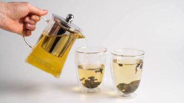 El, şeffaf bir çaydanlıktan cam bardaklara beyaz çay döküyor. Beyaz arka planda buğulanmış beyaz çay.