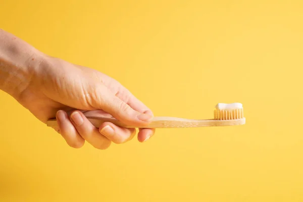 Hand holding bamboo toothbrush on yellow. Holding wooden eco toothbrush. Natural toothpaste on a bamboo toothbrush — Stock Photo, Image