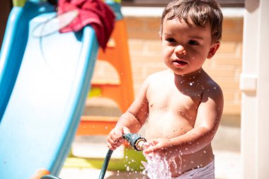Mutlu küçük çocuk arka bahçede su hortumuyla oynuyor.