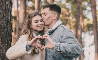 Çam ormanlarında öpüşen ve ellerinden kalp yapan bir çift.