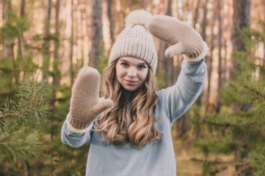 Çam ormanında bir kız şapka ve kalın bir süveter giyer, ellerini eldivenle gösterir ve gülümser.