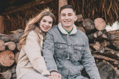 Bir çift, çardaktaki bir bankta yakacak odunların arka planında el ele tutuşup gülümsüyor ve çerçeveye bakıyor.