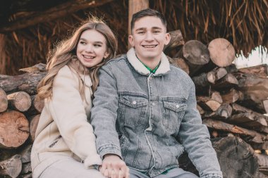 Bir çift, çardaktaki bir bankta yakacak odunların arka planında el ele tutuşup gülümsüyor ve çerçeveye bakıyor.