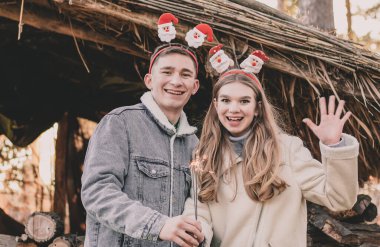 Kafalarında yeni yıl çemberleri olan bir çift ellerinde maytap tutuyorlar ve yakacak odundan yapılmış bir çardağın arka planına gülümsüyorlar.