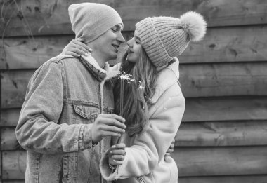 Kışlık giysiler giymiş bir çift ellerinde maytapla ahşap arka planda siyah beyaz bir fotoğraf tutuyorlar.