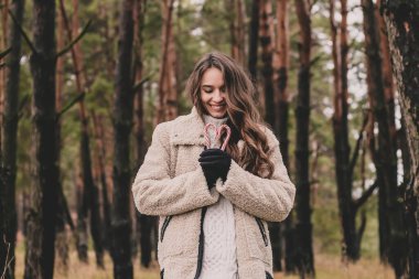 Uzun kıvırcık saçlı, uzun saçlı, güzel bir kadın model elinde kalp şeklinde bir Noel şekeri ve çam ormanındayken gülümsüyor.