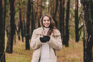 Uzun kıvırcık saçlı, uzun saçlı, güzel bir kadın model elinde kalp şeklinde bir Noel şekeri ve çam ormanındayken gülümsüyor.