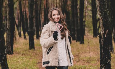 Uzun kıvırcık saçlı, uzun saçlı, güzel bir kadın model elinde kalp şeklinde bir Noel şekeri ve çam ormanındayken gülümsüyor.