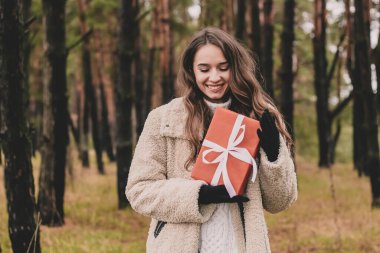 Uzun kıvırcık saçlı, Noel hediyesi tutan ve çam ormanında gülümseyen güzel bir kadın modeli.