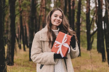 Uzun kıvırcık saçlı, Noel hediyesi tutan ve çam ormanında gülümseyen güzel bir kadın modeli.