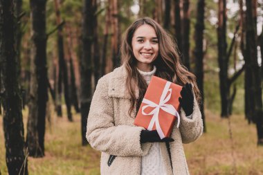 Uzun kıvırcık saçlı, Noel hediyesi tutan ve çam ormanında gülümseyen güzel bir kadın modeli.