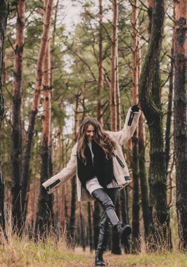 Uzun siyah saçlı ince bir kadın kürk manto, kazak ve deri pantolon giyiyor ve kışın çam ormanlarında bir patika boyunca yürüyor.