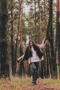 Uzun siyah saçlı ince bir kadın kürk manto, kazak ve deri pantolon giyiyor ve kışın çam ormanlarında bir patika boyunca yürüyor.