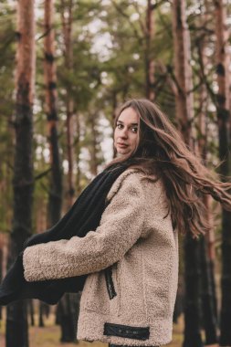Rüzgarda uçan uzun siyah saçlı, model görünümlü bir kadın elinde bir eşarp tutuyor ve çam ormanında gülümsüyor.