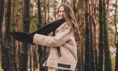 Uzun siyah saçlı ince bir kadın kürk manto giyiyor, bir kazak ve deri pantolon kışın bir çam ormanının arka planında yürür.