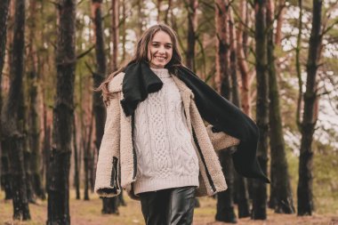 Uzun siyah saçlı ince bir kadın kürk manto giyiyor, bir kazak ve deri pantolon kışın bir çam ormanının arka planında yürür.