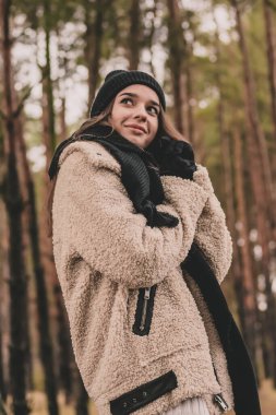 Uzun siyah saçlı ince bir kadın kürk manto giyiyor, bir kazak ve deri pantolon kışın bir çam ormanının arka planında yürür.