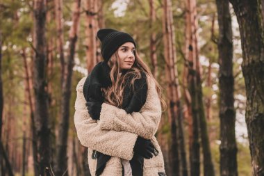 Uzun siyah saçlı ince bir kadın kürk manto giyiyor, bir kazak ve deri pantolon kışın bir çam ormanının arka planında yürür.