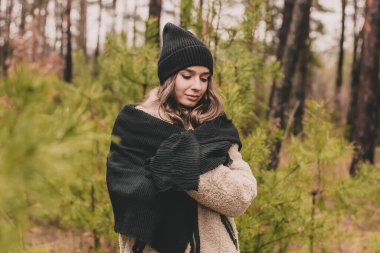 Süveter şapkalı ve kürklü uzun kıvırcık saçlı, manken görünümlü güzel bir kadın çam ormanındaki Noel ağaçlarının arka planında duruyor.