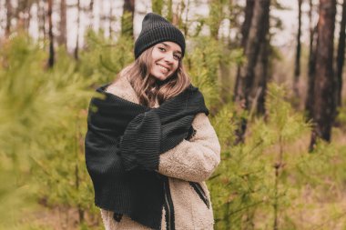 Süveter şapkalı ve kürklü uzun kıvırcık saçlı, manken görünümlü güzel bir kadın çam ormanındaki Noel ağaçlarının arka planında duruyor.