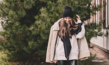 Uzun kıvırcık saçlı, süveter şapkalı ve kürklü güzel bir kadın Noel ağacının arka planında ve evinde duruyor.