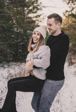 Bir adam, karlı bir ormanın arka planında bir kadını kollarının arasına alır.