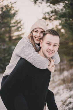 Manken görünümlü bir kadın bir erkeğin sırtına oturur ve karlı bir ormanın arka planında boynuna sarılır.